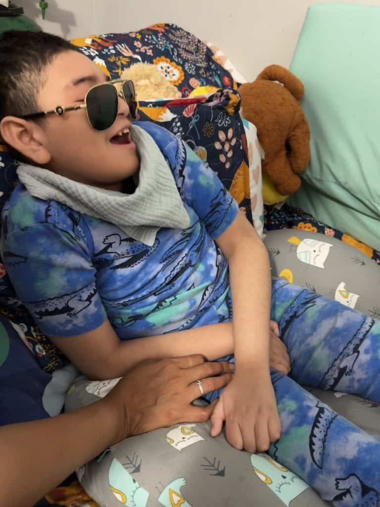 Lucas, a medically complex child with special needs relaxes in his adjustable bed wearing aviator sunglasses. HIs bib looks like a scarf.